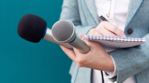 Eine Person hält zwei Mikrofone in der einen Hand einen Notzblock und Stift in der anderen Hand